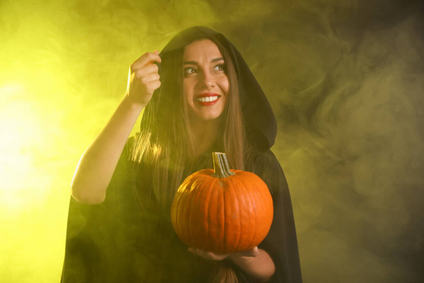 Young woman wearing witch costume with pumpkin in smoke cloud on dark background. Halloween party - Foto, afbeelding