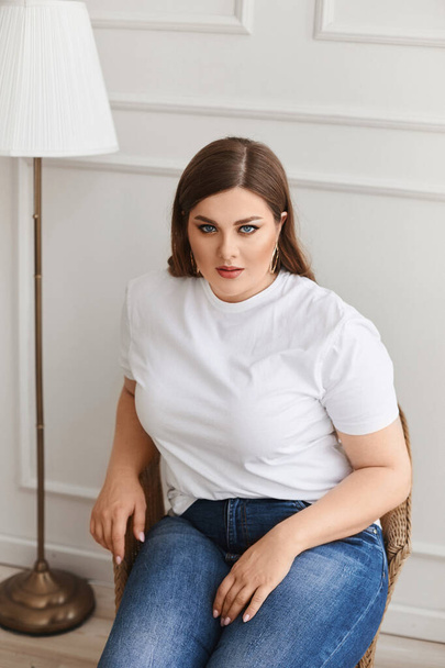 Plus size model girl in jeans and blank white t-shirt sits in the armchair in the living room. XXL fashion. - Fotoğraf, Görsel