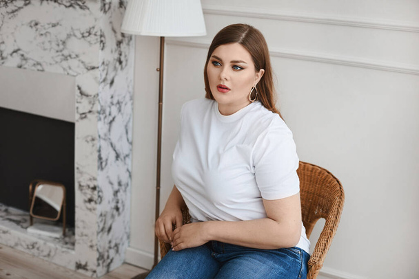 Plus size model girl in jeans and blank white t-shirt sits in the armchair in the living room. XXL fashion. - Fotografie, Obrázek