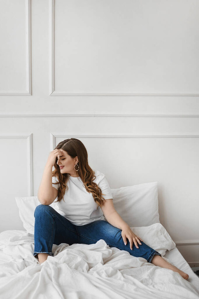 Plus-size model girl in jeans and blank white t-shirt sitting on the bed. XXL fashion. - Valokuva, kuva