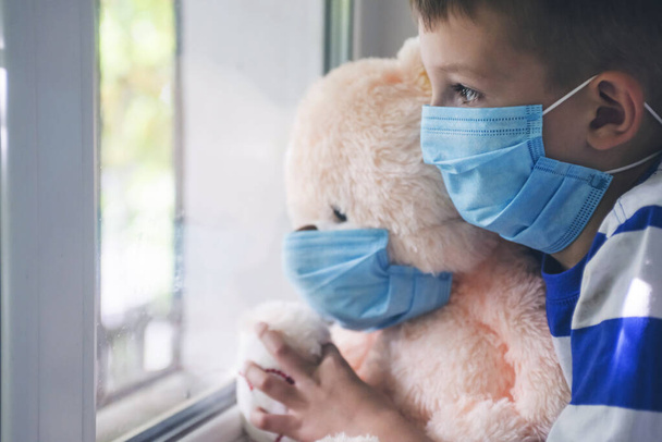 Sad child and his teddy bear both in protective medical masks looks out window. Virus protection, coronavirus pandemic. Coronavirus covid-2019. - Foto, Bild