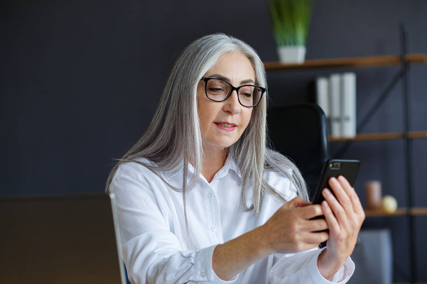 Senior grey-haired beautiful woman using smartphone and smiling. Happy businesswoman using mobile phone apps, texting message, browsing internet, shopping online. Mature people with mobile devices - 写真・画像