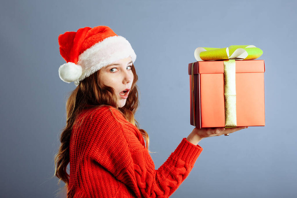 Hermosa dama europea en Santa sombrero está sosteniendo regalos, mirando a la cámara y sonriendo, aislado sobre fondo gris. - Foto, Imagen