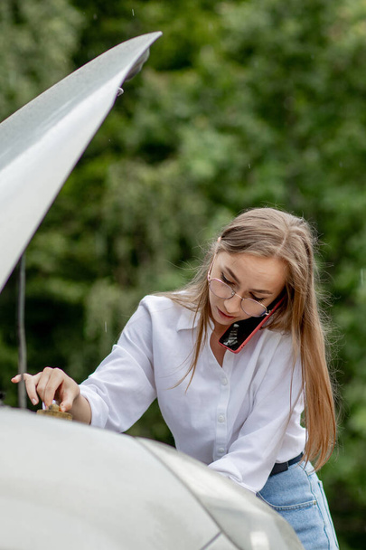 Young woman standing near broken down car with popped up hood having trouble with her vehicle. Waiting for help tow truck or technical support. A woman calls the service center - Фото, зображення