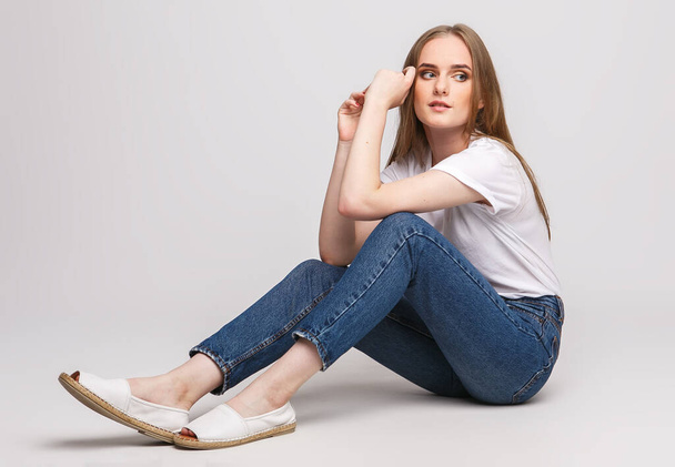 молода красива жінка в білій футболці і джинсах на білому тлі. Молода жінка сидить на білому тлі. Фото концепція реклами одягу та взуття
. - Фото, зображення