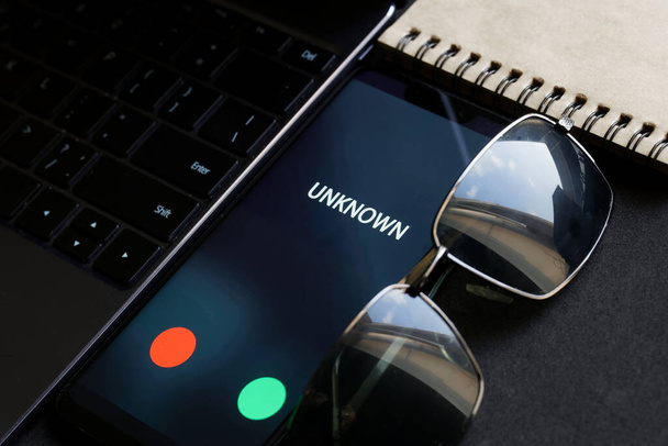Ένα μαύρο smartphone με μια άγνωστη κλήση βρίσκεται δίπλα στο φορητό υπολογιστή με γυαλιά ηλίου και ένα σημειωματάριο. Το πρόβλημα με το σκοτεινό δίκτυο, το χακάρισμα και την κατασκοπεία. Προστασία των προσωπικών πληροφοριών. Ρηχό βάθος πεδίου - Φωτογραφία, εικόνα