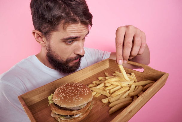 Férfi egy tálca étel a kezében hamburger sült krumpli és gyorsétterem kalória rózsaszín háttér portré közelkép - Fotó, kép