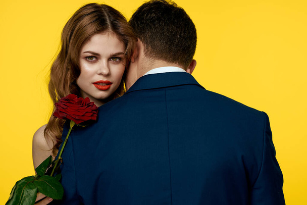 A romantic man hugs a woman in a red dress with a rose in his hand on a yellow background - Фото, зображення