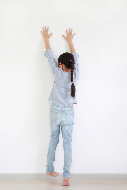 Asian little child girl raise hand up and climbing posture at white wall of room. Back view. - Фото, зображення