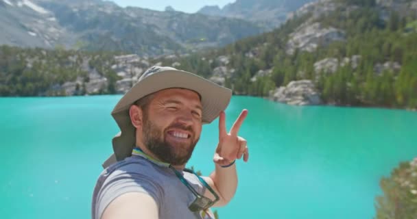 Comical όμορφος άντρας στο καπέλο κυνηγός χρησιμοποιώντας smartphone για να δείξει τη λίμνη και τα βουνά - Πλάνα, βίντεο