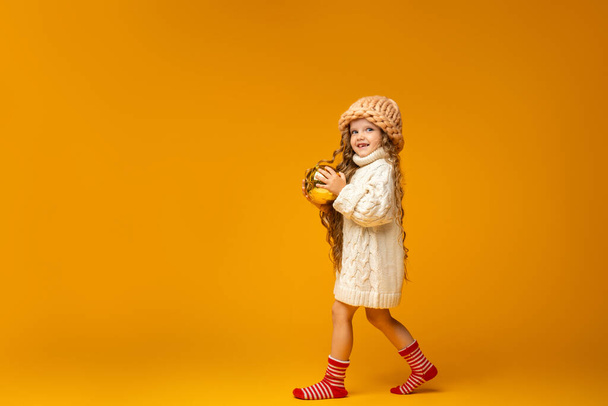 little Caucasian girl 6 years old in knitted clothes in Christmas socks and with large red Christmas ball in her hands is walking on yellow background in Studio. Preparing for new year. Copy space. - Foto, afbeelding