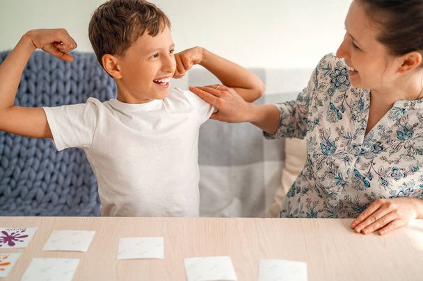 Mom and son 8 years old play flashcards. Boy wins and shows his strength, the muscles in his arms. The woman smiles. Winner, success, communication between mother and son. Parenthood. - Foto, Imagen