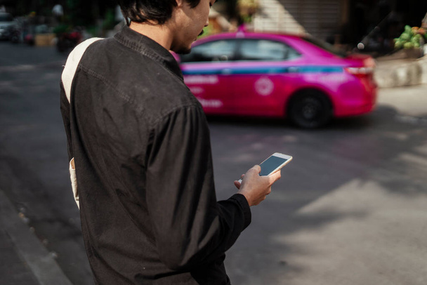 Μαύρα μαλλιά άνθρωπος στέκεται στο μονοπάτι και να χρησιμοποιήσετε το smartphone του, ενώ η κίνηση ταξί στο δρόμο. - Φωτογραφία, εικόνα