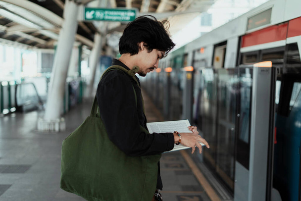 Μαύρα μαλλιά τύπος στέκεται ήρεμα και να διαβάσετε το βιβλίο στο σταθμό Skytrain, ενώ άλλοι άνθρωποι προσπαθούν να μπουν μέσα στο τρένο. - Φωτογραφία, εικόνα