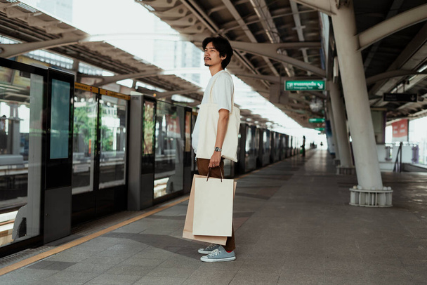 Muž v bílém tričku drží papírovou nákupní tašku a čeká, až se Skytrain vrátí domů.. - Fotografie, Obrázek