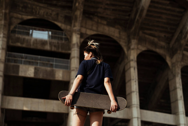 Woman in dark blue polo shirt hold the longboard at her butt standing in front of pretended colosseum building. - 写真・画像