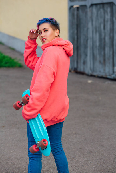 Stylish young woman with short colored hair holding blue plastic skateboard in hand. Youth concept. - Foto, Imagen