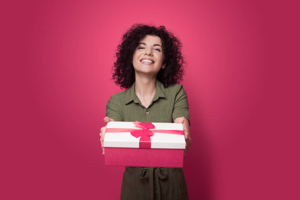 Brunette woman with curly hair is giving a present at camera being very happy and generous on a red studio wall during holidays - Foto, Imagem