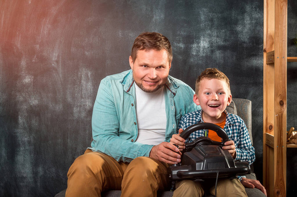 Isä ja poika pelaavat videopelejä sohvalla. Onnellinen isä ja poika pyöräkonsolilla pelatessaan peliä kotona. Riippuvuus ja teknologia, perheen ja ihmisten käsite. - Valokuva, kuva