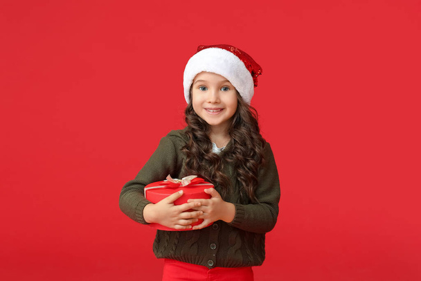 Cute little girl in Santa hat and with gift on color background - Foto, Imagem