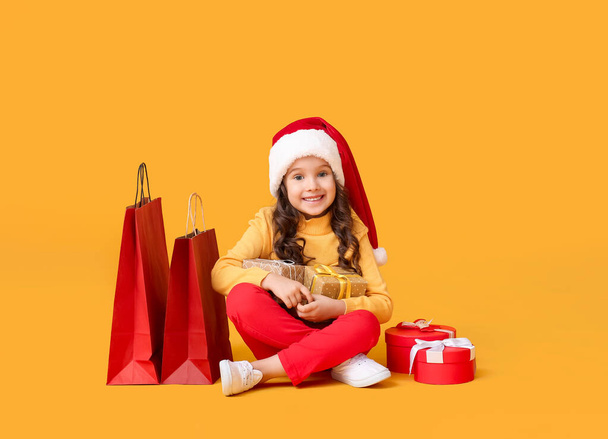 Χαριτωμένο κοριτσάκι σε Σάντα καπέλο, με κουτιά δώρων και τσάντες ψώνια στο φόντο χρώμα - Φωτογραφία, εικόνα