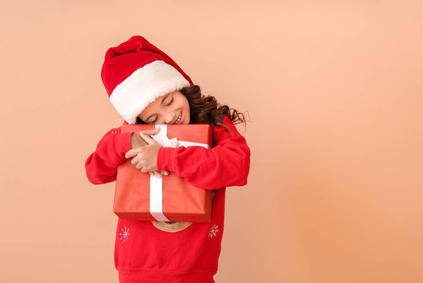 Nettes kleines Mädchen mit Weihnachtsmütze und mit Geschenk auf farbigem Hintergrund - Foto, Bild