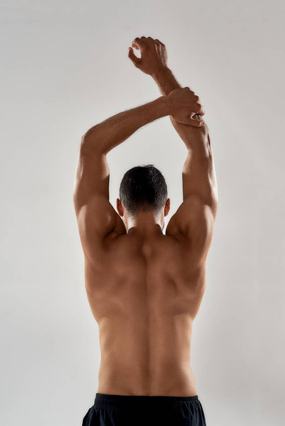 Rear view of muscular man with naked torso raising his arms above head isolated over grey background, posing in studio - Fotografie, Obrázek