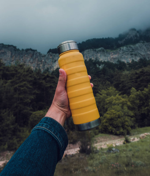 man hand met een stalen gele thermo fles op natuur achtergrond. - Foto, afbeelding