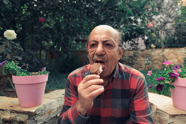 Primer plano Retrato del hombre mayor comiendo helado. Anciano comiendo helado en palo en el patio trasero. - Foto, imagen