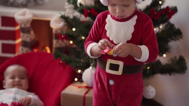 Vánoce, zima, Nový rok, Celebration, rodina, Dětství koncept - šťastný chlapeček v Santa klobouku a vánoční oblečení.otevírá cukroví a jí. novorozeně dítě po krbu hrát. Sourozenci se baví na dovolené - Záběry, video