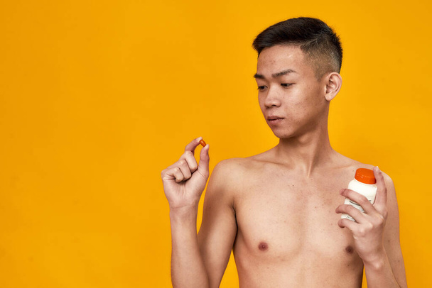Portrait of shirtless young asian man looking at red capsule, holding jar of pills isolated over yellow background. Health care, treatment concept - Fotoğraf, Görsel