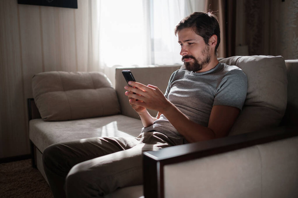 Egy vonzó, mosolygós, fiatal, szakállas férfi portréja, aki alkalmi ruhákat visel, és a nappaliban ül a kanapén. hipster srác használja a közösségi média app messenger, szörfözés web telefon beltéri - Fotó, kép
