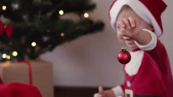 クリスマス、冬、新年、お祝い、家族、子供の概念-サンタの帽子とクリスマス服の幸せな面白い子供の男の子は遊び、赤いボールで笑顔は休日にクリスマスツリーを飾る. - 映像、動画