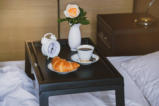 Happy morning. Breakfast in bed on tray table with coffee and croissant and rose in vase, wooden interior, white bed. - Фото, зображення