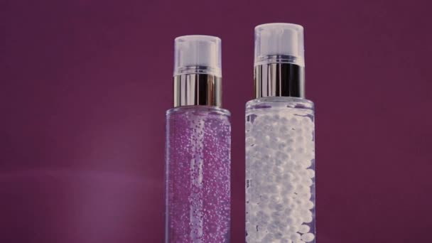 Meikkipohja ja seerumigeeli luksusihonhoitotuotteina ja kiiltävinä valosoihdutuksina violetilla pohjalla, ihonhoitorutiinina kasvojen kosmetiikassa ja kauneusmerkkinä - Materiaali, video