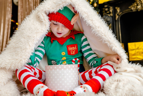 Un niño lindo vestido como un elfo abre un regalo de Navidad mientras se esconde detrás de una manta esponjosa en una casa de Navidad decorada. - Foto, imagen