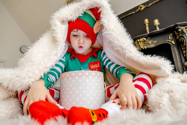 A cute little boy dressed as an elf opens a Christmas present while hiding behind a fluffy blanket in a decorated Christmas house. - Photo, Image