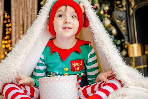 A cute little boy dressed as an elf opens a Christmas present while hiding behind a fluffy blanket in a decorated Christmas house. - Photo, Image