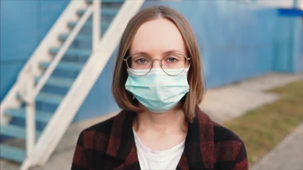 Menina em óculos de moda, máscara protetora médica e casaco olha para a câmera.  - Filmagem, Vídeo