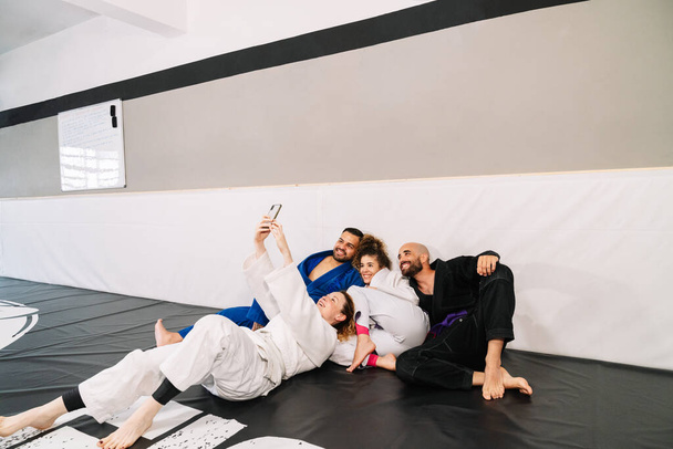 Portrait de quatre compagnons d'arts martiaux comme le judo en utilisant le kimono assis sur le tapis du sol et appuyé sur le mur de la salle de gym, ce qui rend un selfie avec le mobile très heureux après un entraînement - Photo, image