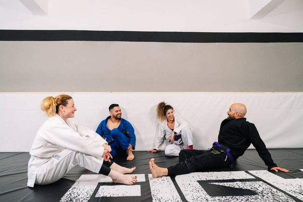 Ritratto di quattro partner di arti marziali come il judo che usano il kimono seduto sul tappetino e appoggiato al muro della palestra, che parlano e ridono dopo un allenamento - Foto, immagini