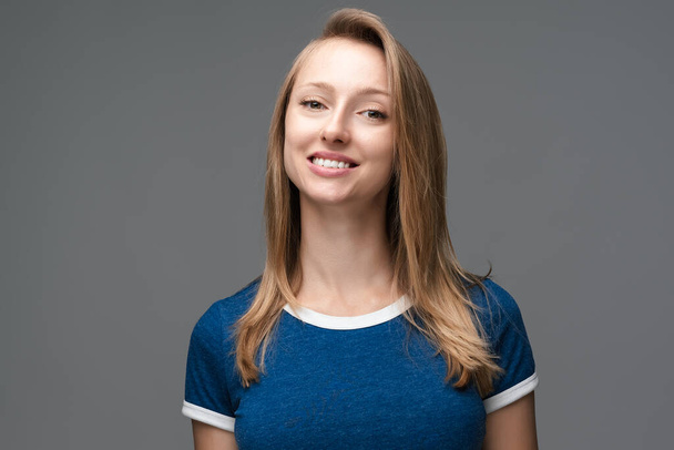 Studio shot of pretty female with blonde straight hair, broad smile, wears blue t shirt, tilts head and looks with joy. Human emotions, facial expression concept. - Photo, Image