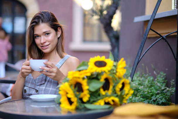 young stylish woman, fashion sunglasses, sitting in cafe, holding drinking cup cappuccino, enjoying, sunflowers, happy stret, city street, europe vacation, romantic dinner, sunny - Foto, Bild