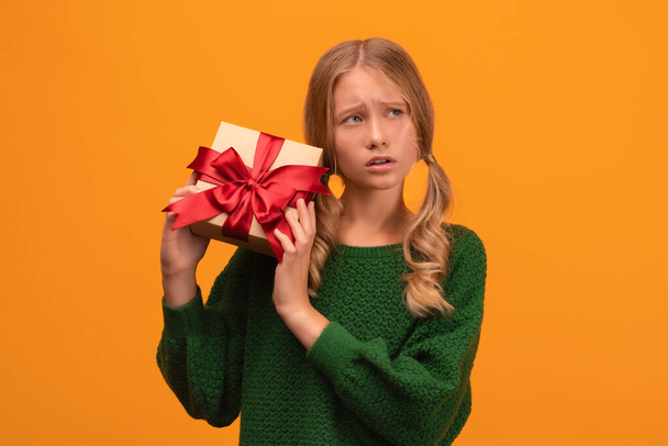 Afbeelding van charmant blond meisje 12-14 jaar oud in warm groene trui met geschenkdoos met rode strik. Studio schot, gele achtergrond, geïsoleerd. Nieuwjaar Vrouwendag verjaardagsvakantie concept - Foto, afbeelding
