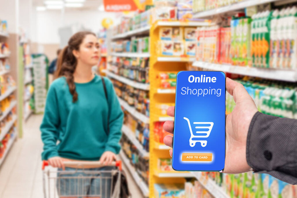 De hand van een persoon houdt een mobiele telefoon vast, met een winkelmandje op het scherm. Op de achtergrond duwt een vrouw een boodschappenkarretje in een supermarkt, in een waas. Het concept van online winkelen. - Foto, afbeelding