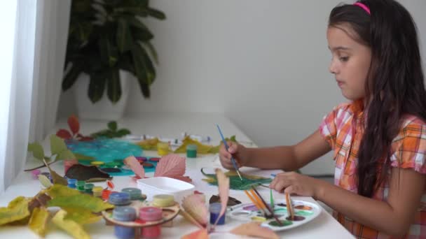 Close-up van het kleine meisje dat aan tafel zit en de bladeren tekent met penseel en aquarellen. klein meisje schilderen gevallen bladeren thuis. herfst - Video