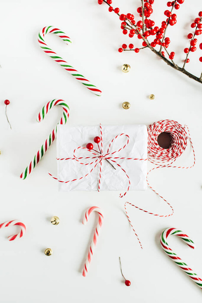 Karácsonyi ajándékdoboz ünnepi cukorkával, piros bogyós ág, karácsonyi kötél tekercs fehér asztalon. Karácsony, újévi kompozíció. Lapos fekvésű, felülnézet - Fotó, kép
