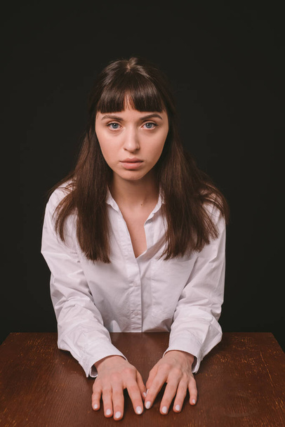 Studio portret van een mooie brunette vrouw in een wit shirt, zitten aan de tafel, tegen een effen zwarte achtergrond, serieus kijken naar de camera - Foto, afbeelding
