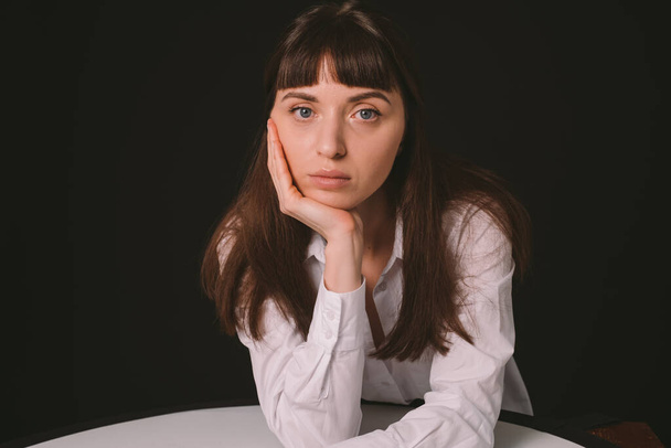 Studio portrait of a pretty brunette woman in a white shirt, sitting at the table, against a plain black background, looking down - Photo, Image