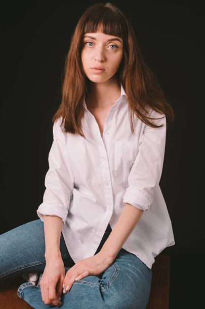 Studio portrait of a pretty brunette woman in a white shirt, sitting against a plain black background, looking at camera - Foto, Imagem